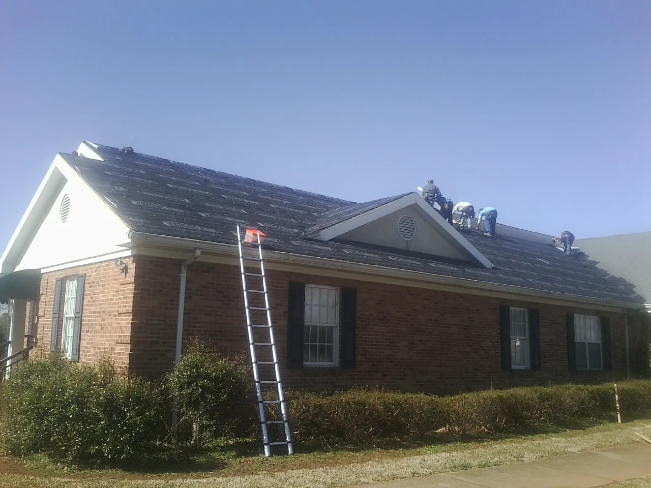 Crew tearing shingles off a brick ranch roof with a ladder set — Metal Roof Repair work by our crew in Alton Bay