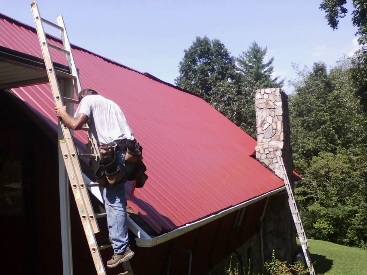 Roofer on a ladder beside a red metal roof and stone chimney — Soffit & Fascia Repair by Roofing Brothers in Alton Bay